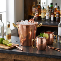 A smooth copper ice bucket with gold handles and a spoon, filled with ice alongside matching copper mugs.