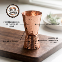 A copper jigger with hammered detail, shown on a wooden board with callout labels and a 'Solid Copper' badge. 