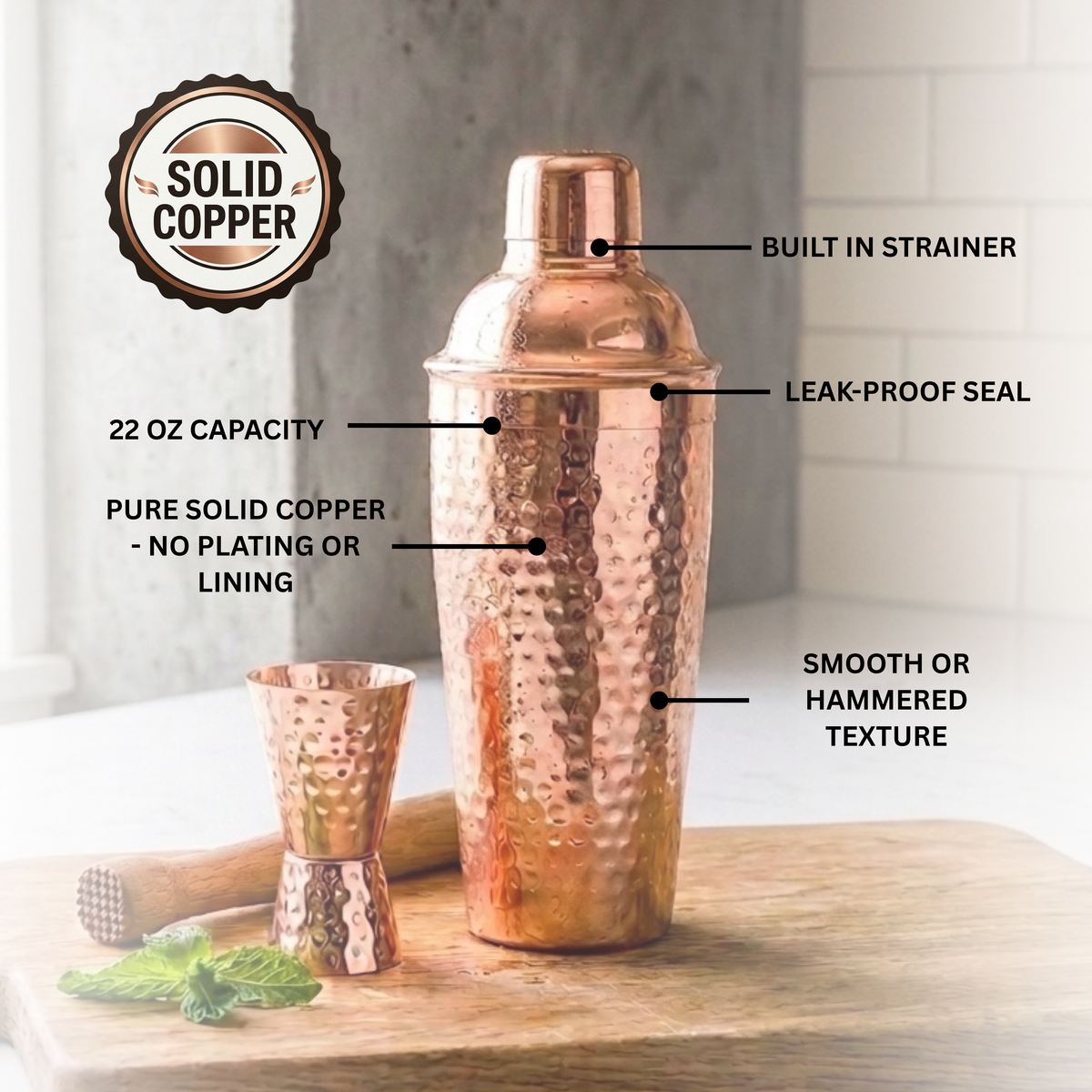 A hammered solid copper cocktail shaker and matching jigger on a wooden surface with callout  text pointing out features like built-in strainer, leak-proof seal, 22 oz capacity, pure solid copper, and hammered texture.