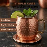 A hammered copper Moscow mule mug with a gold-toned handle sits on a matching copper coaster on a dark marble surface. 