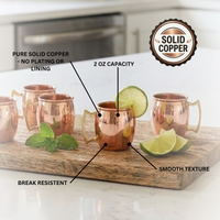 Four mini 2oz copper Moscow mule cups arranged on a wooden board with lime wedges and mint leaves, overlaid with callout labels and a circular badge reading 'Solid Copper.' 