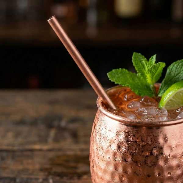 A copper Moscow mule cocktail in a hammered copper mug, filled with ice and garnished with mint leaves and a lime wedge, with a copper straw, on a dark wooden surface.