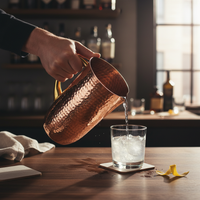 Copper pitcher being used to pour a drink into a glass with ice on a table.