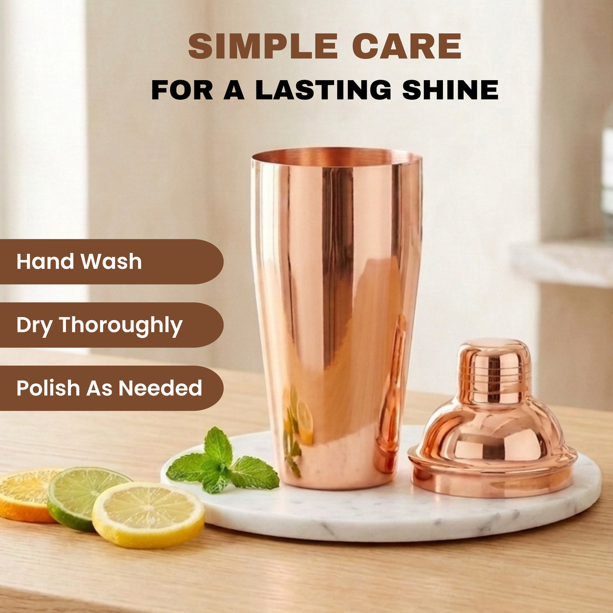 A copper cocktail shaker and matching lid displayed on a white marble tray on a wooden table, with lemon slices, mint leaves, and text reading Hand Wash, Dry Thoroughly and Polish As Needed.