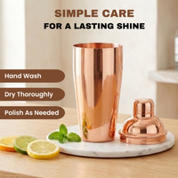 A copper cocktail shaker and matching lid displayed on a white marble tray on a wooden table, with lemon slices, mint leaves, and text reading Hand Wash, Dry Thoroughly and Polish As Needed.