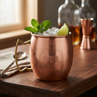 A copper Moscow mule mug with a gold-toned handle and engraved monogram, filled with ice, mint leaves, and a lime wedge on a wooden bar surface with bottles in the background.