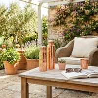 Two sizes of a solid copper water bottle on an outdoor patio with wicker chair, wooden table, and decorative elements.