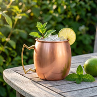 A copper Moscow mule mug with a gold handle sits on a weathered wooden table outdoors, filled with ice, garnished with mint leaves and a lime wheel, with a whole lime nearby and a blurred green garden background.
