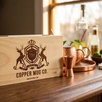 A wooden gift box engraved with the Copper Mug Co. crest and name sits on a wooden table beside copper mugs, a jigger, and limes in a warm, softly lit bar setting.